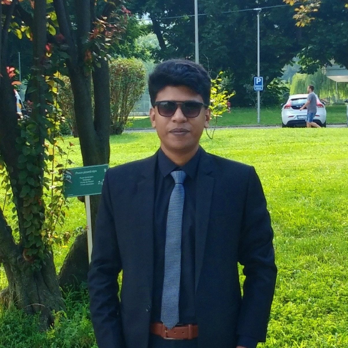 Raihan Khan in a navy suit and striped tie with sunglasses standing in a European park lined with trees.