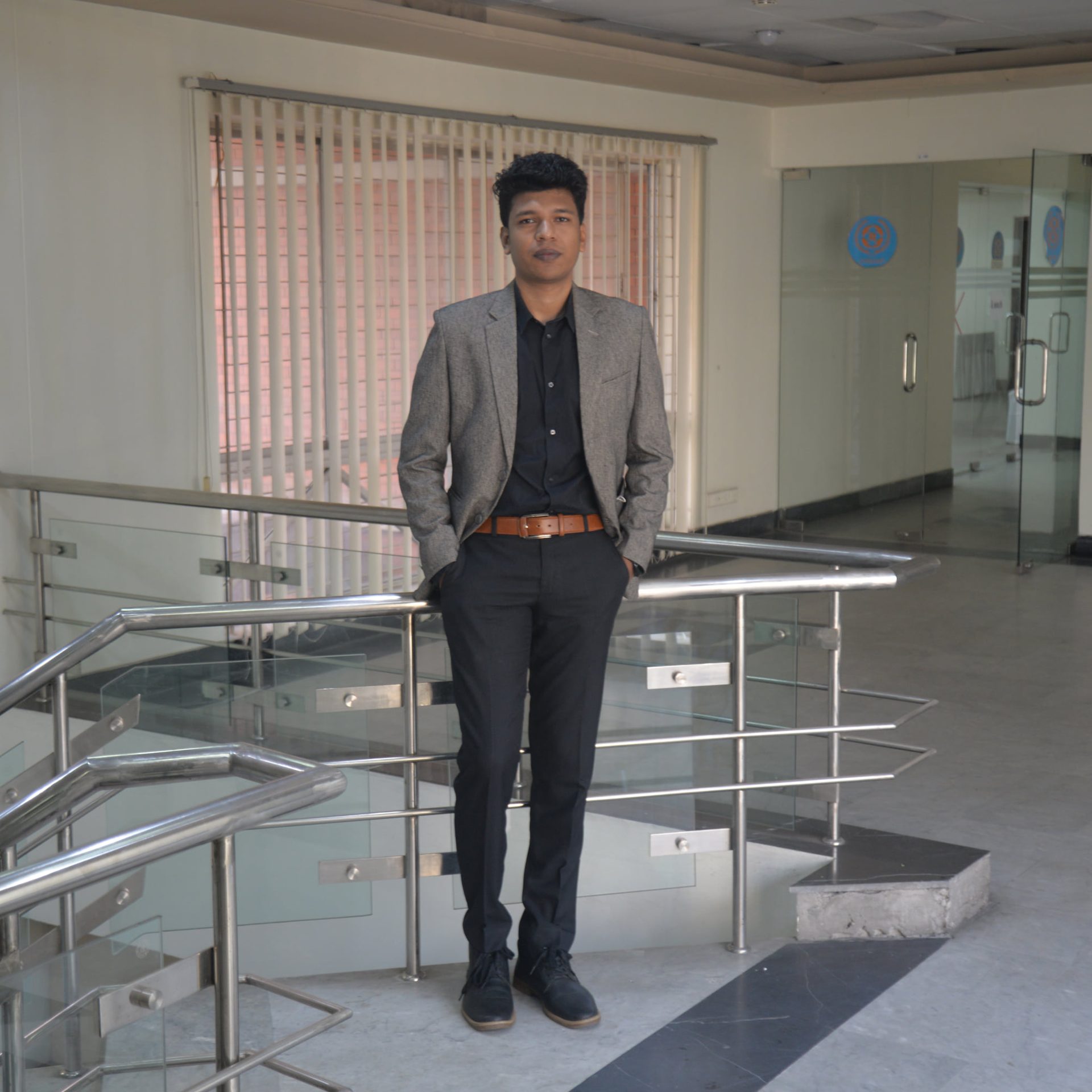 Raihan Khan in a grey blazer standing with hands in pockets near a glass staircase in an office corridor.