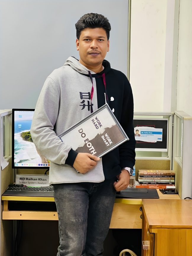 Raihan Khan in a grey and black hoodie holding a Work Hard Work Smart plaque at his office desk.