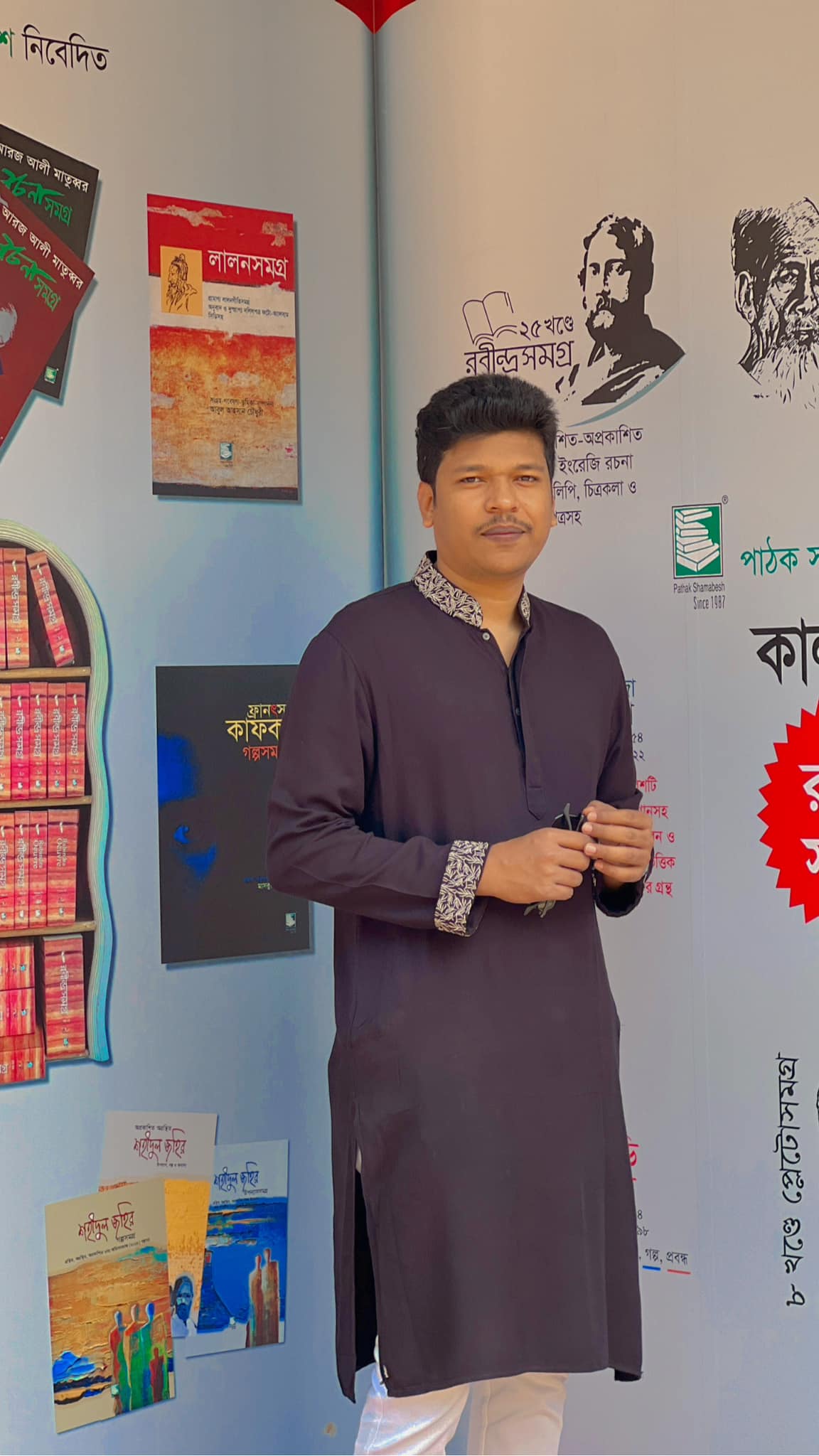 Raihan Khan in a plum kurta standing before a wall of book covers at a literary book fair exhibit.