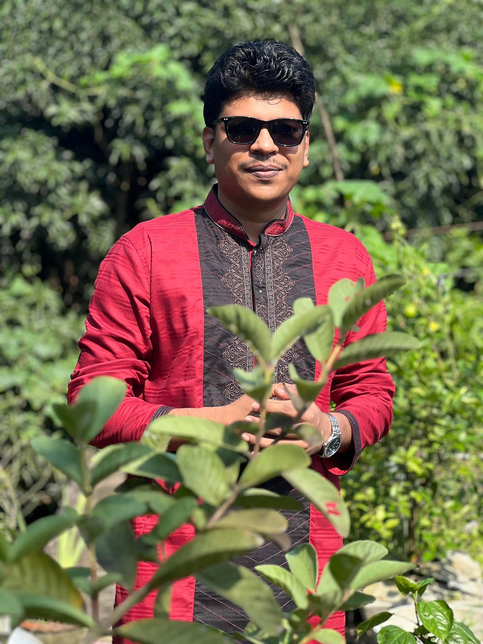 Raihan Khan in a red and black kurta with sunglasses standing behind garden leaves on a sunny day.