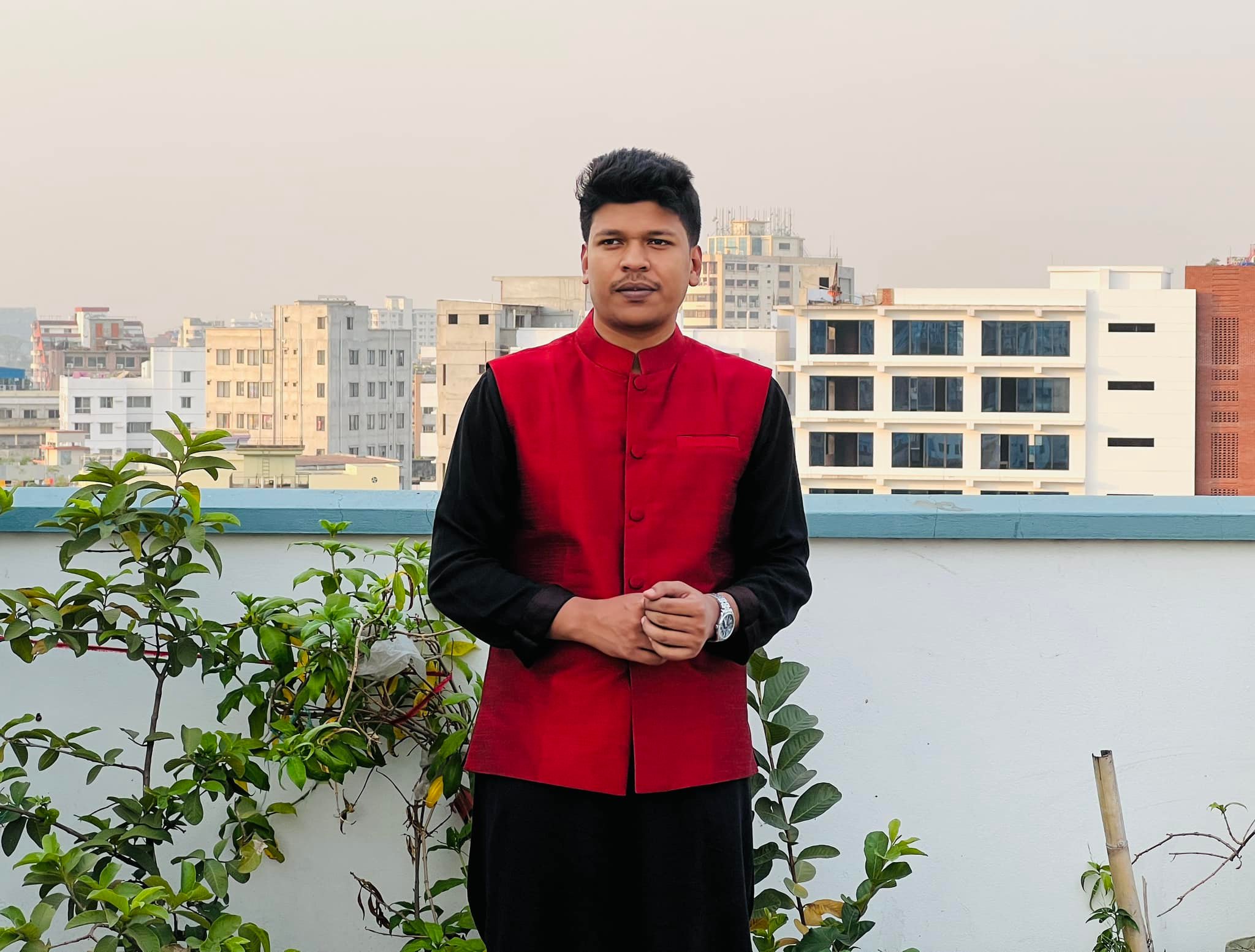 Raihan Khan in a red waistcoat over a black kurta standing on a rooftop with the city skyline behind him.