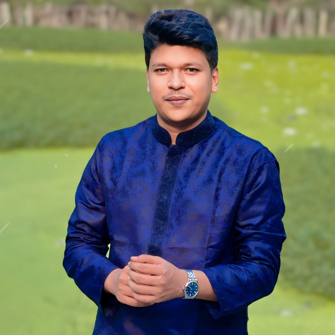 Raihan Khan in an embroidered royal blue kurta posing with clasped hands against a green studio backdrop.