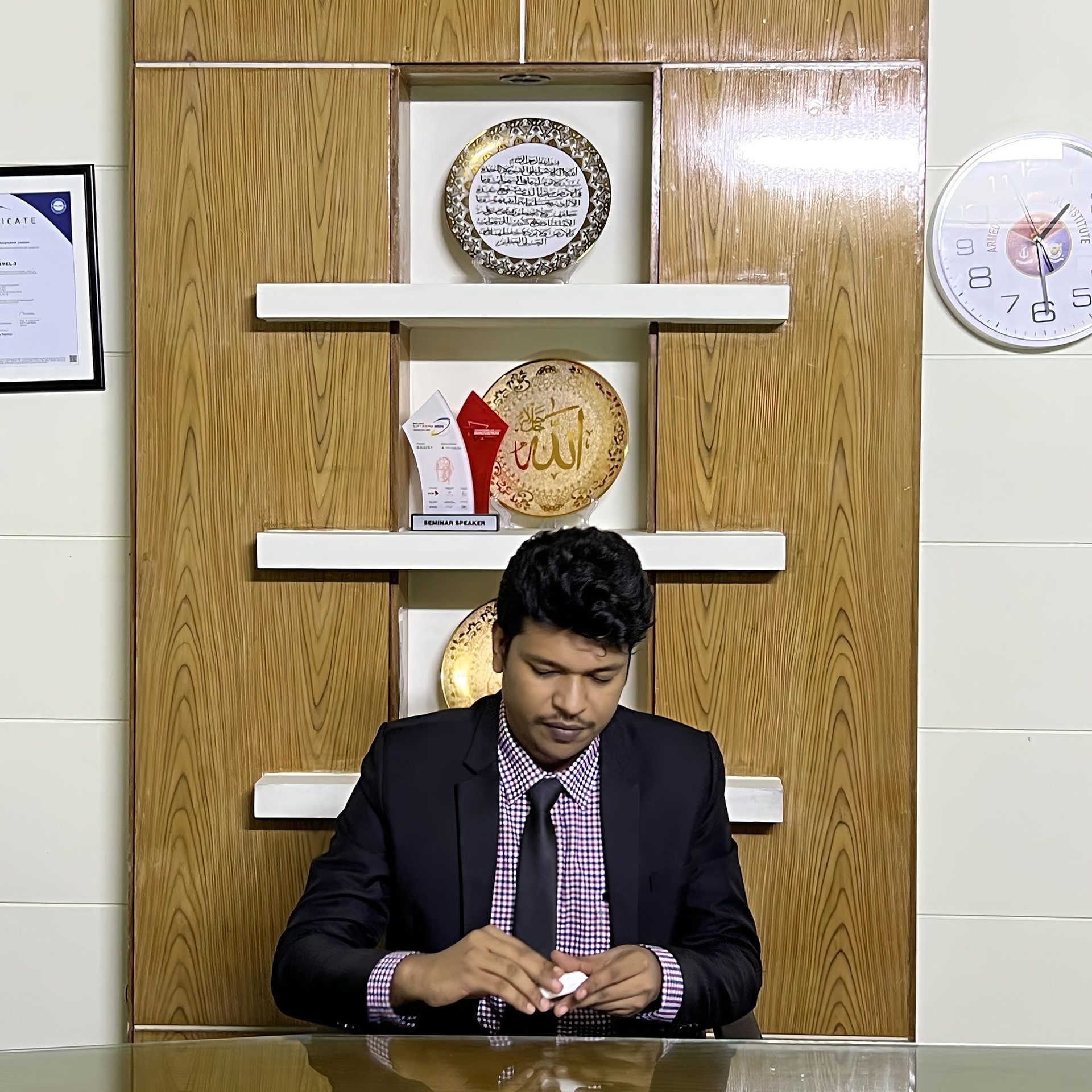 Raihan Khan in a dark suit and checkered shirt seated at a desk beneath a Seminar Speaker award and wall clock.