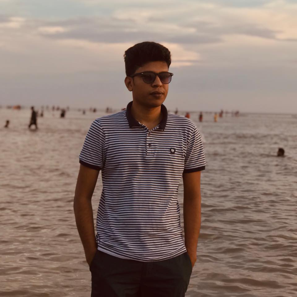 Raihan Khan in a striped polo shirt and sunglasses standing on a beach with the ocean behind him.
