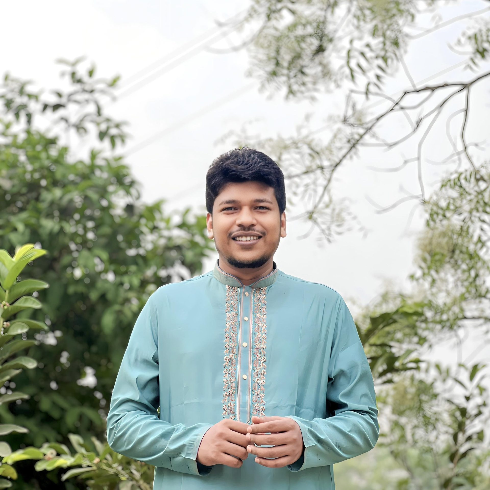 Raihan Khan in an embroidered teal kurta smiling broadly beneath tree branches outdoors.