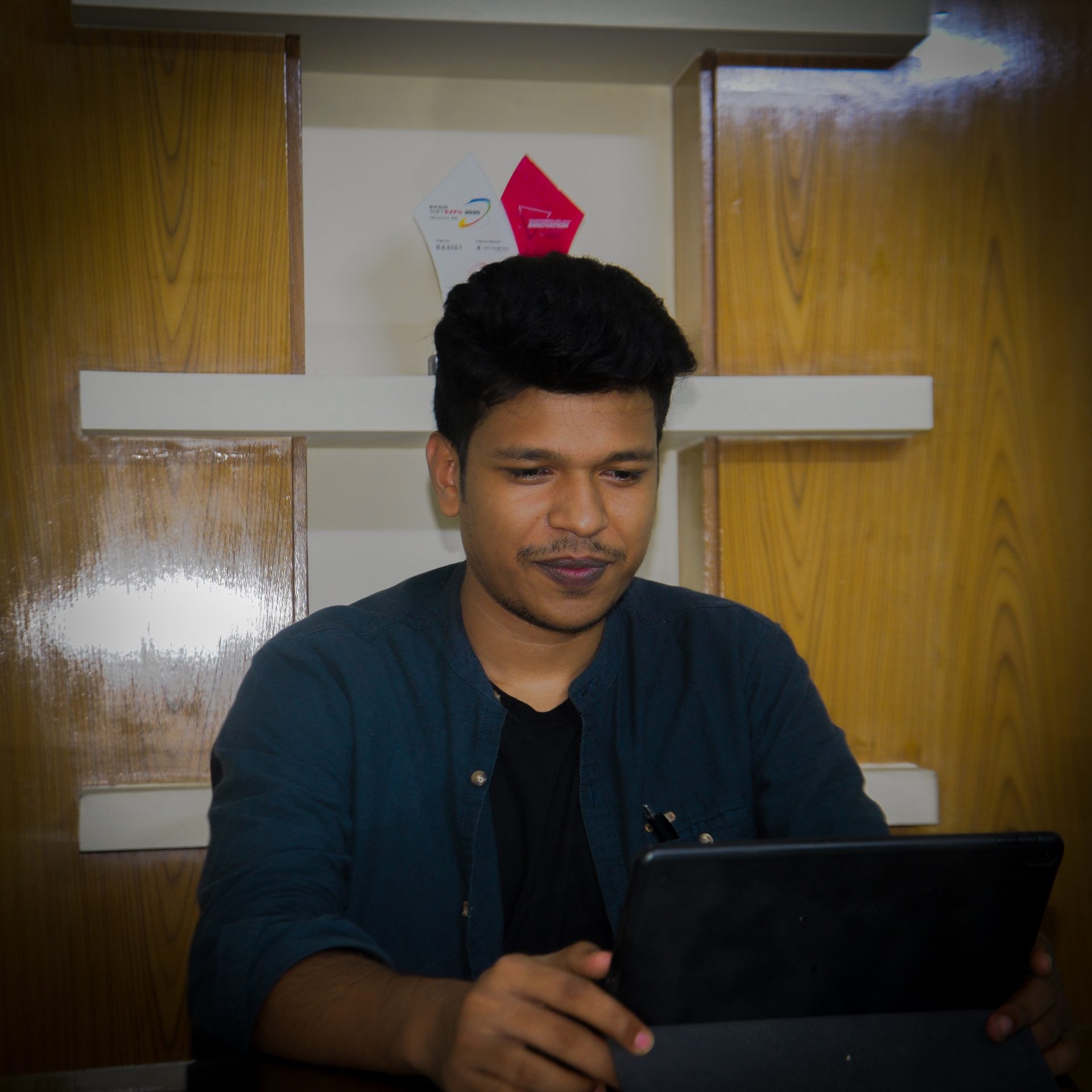 Raihan Khan in a teal shirt looking down at a tablet with award certificates displayed on the shelf behind him.