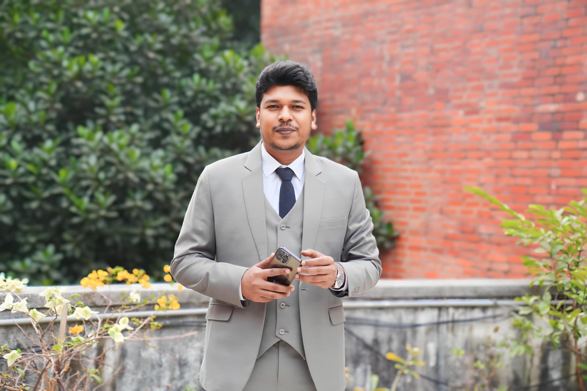 Raihan Khan in a grey three-piece suit and tie holding his phone in front of a red brick wall.