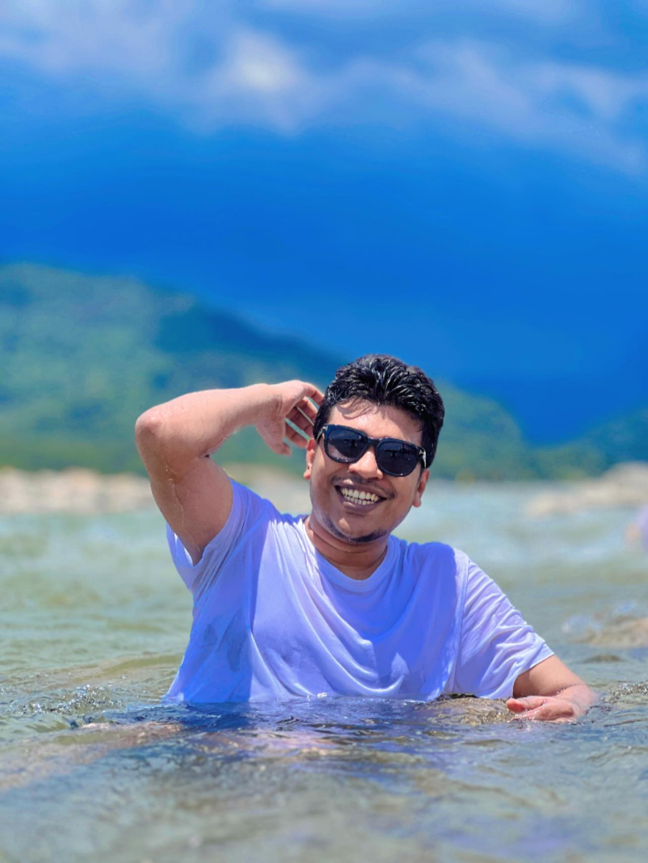 Raihan Khan laughing while wading in the sea in a wet white t-shirt with mountains in the distance.