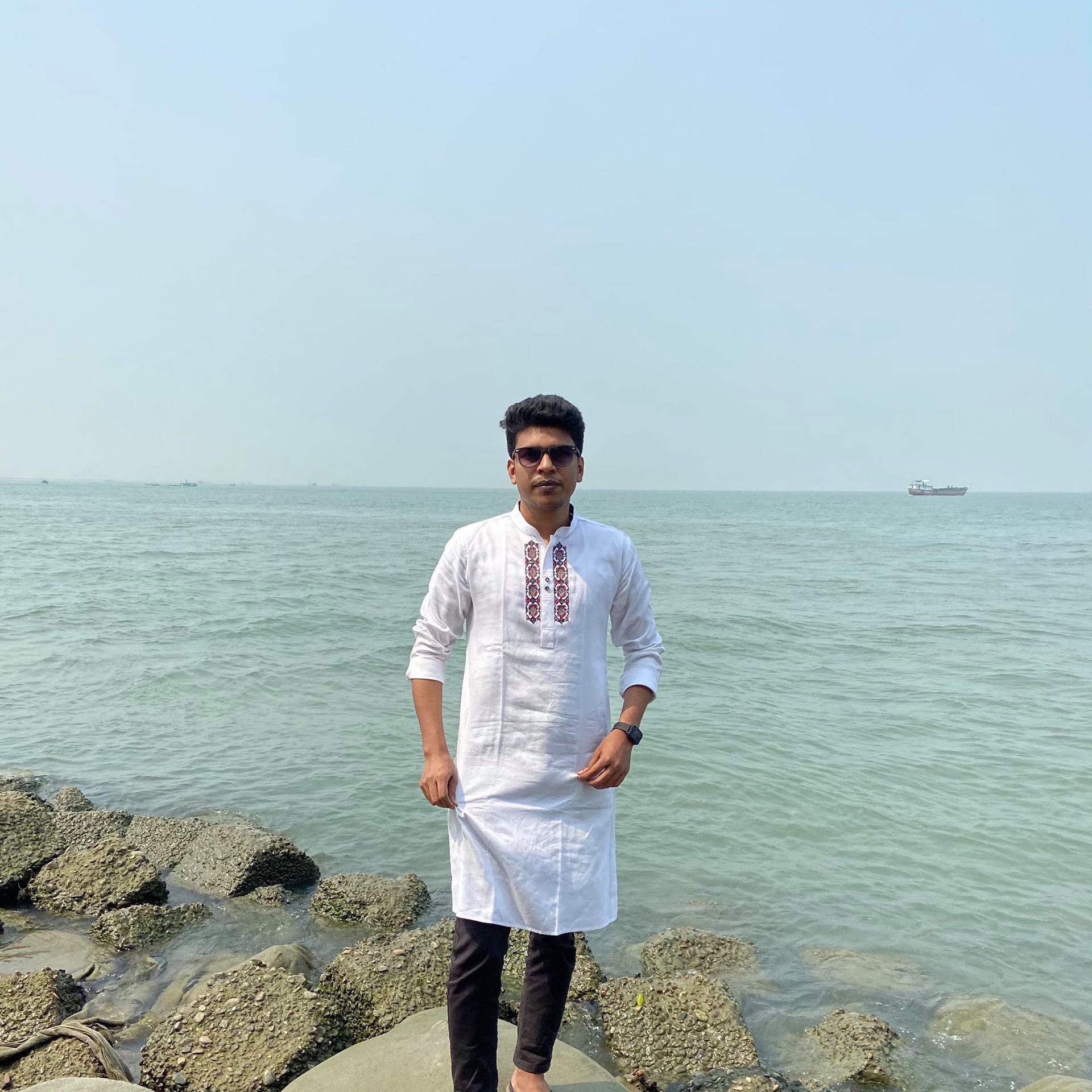 Raihan Khan in an embroidered white kurta and sunglasses standing on a rocky shoreline with ships out at sea.