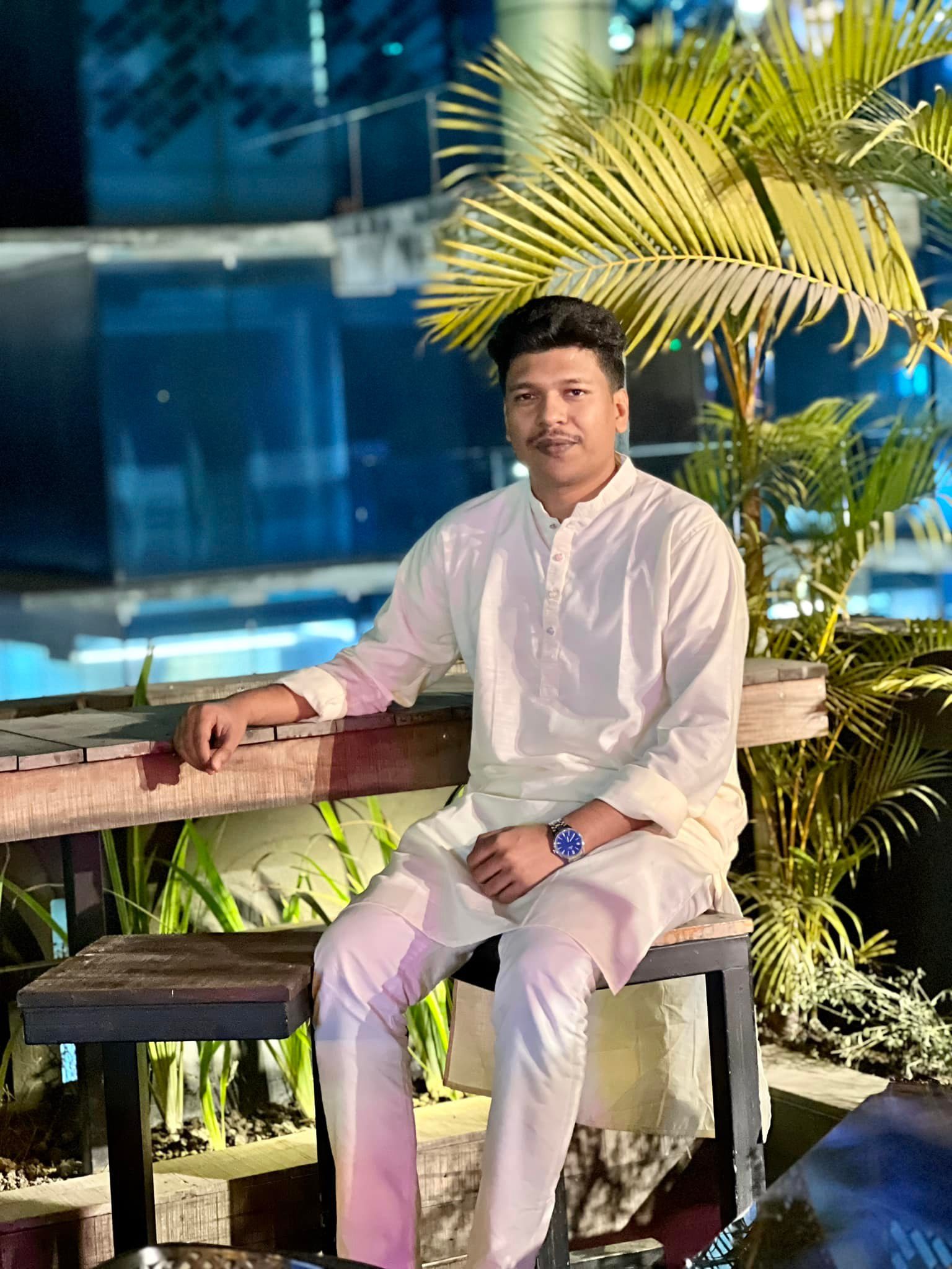 Raihan Khan in a white kurta sitting on a wooden bench beside a palm tree on a rooftop at night.