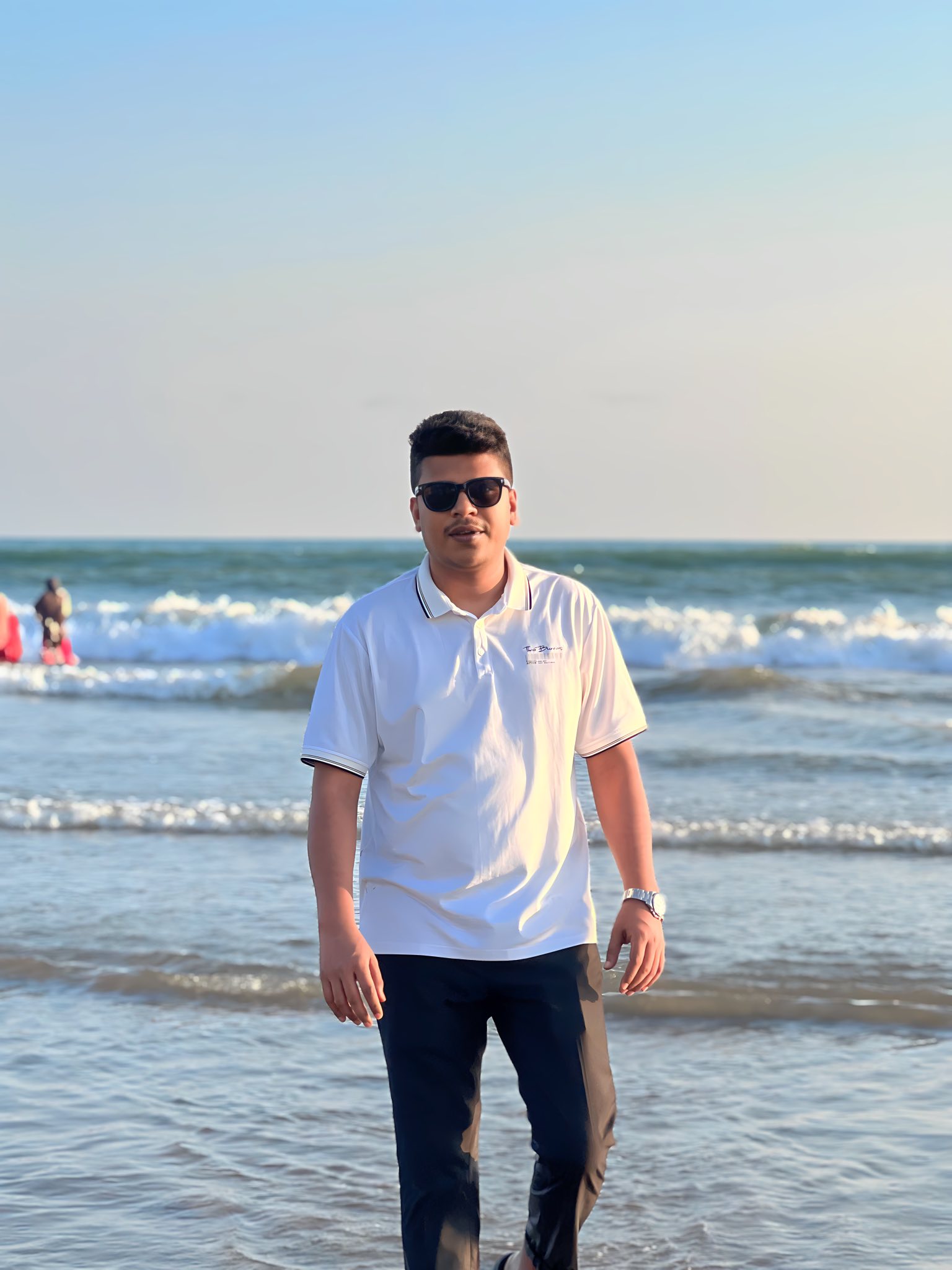 Raihan Khan in a white polo shirt and sunglasses walking along a beach with waves rolling in behind him.