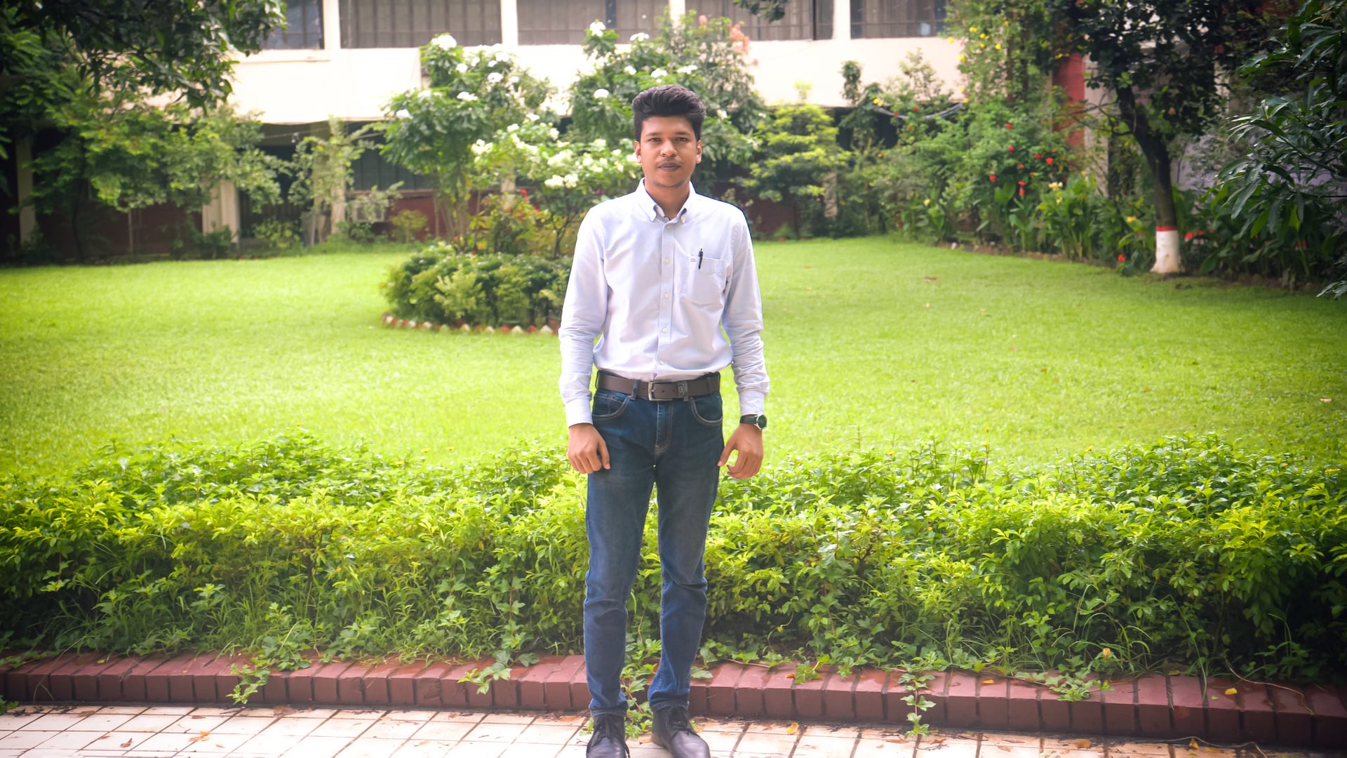 Raihan Khan in a white shirt and jeans standing full-length on a garden path lined with green lawns.