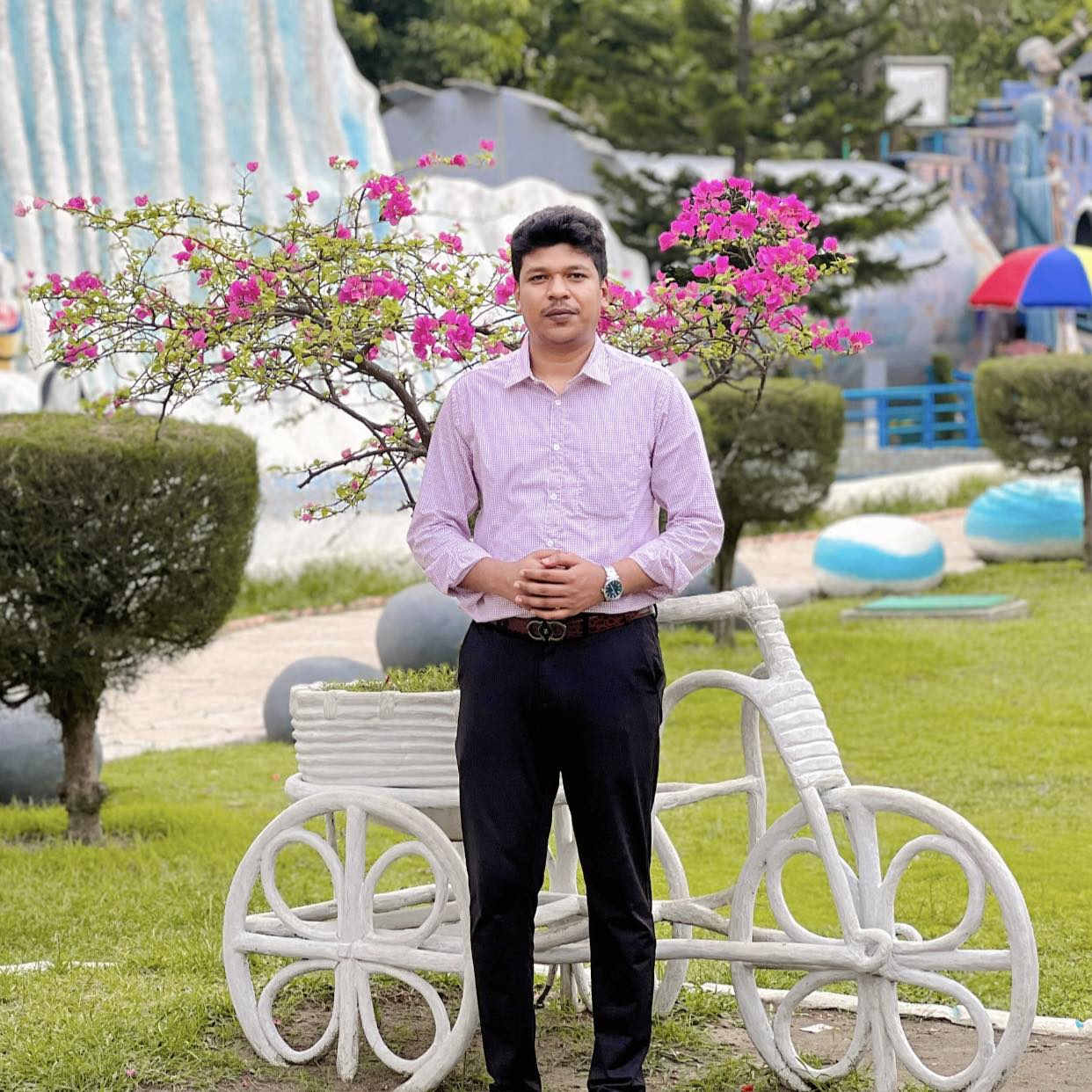 Raihan Khan standing in an amusement park surrounded by bougainvillea flowers and a stone bicycle statue.