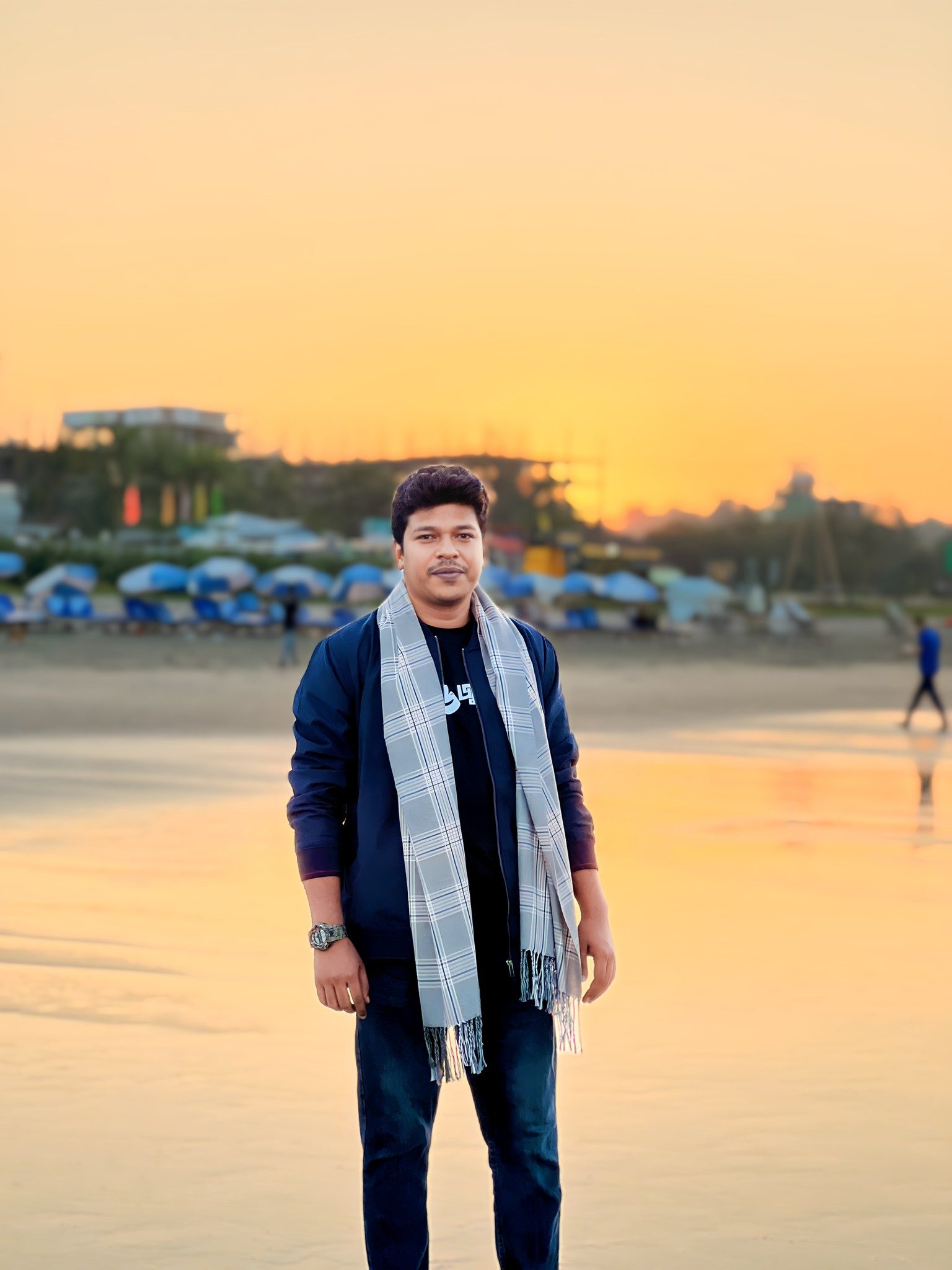 Raihan Khan poses on a beach at golden hour, wrapped in a checkered scarf as the sun sets over the shoreline.