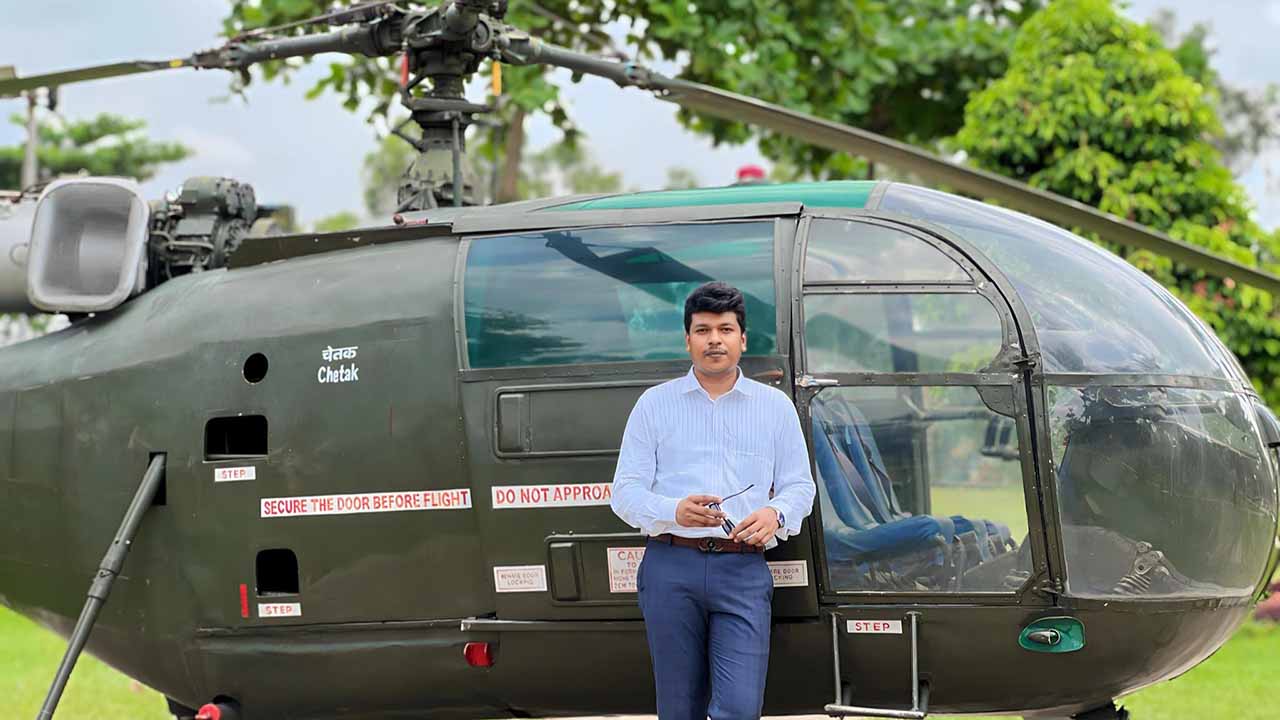 Raihan Khan stands beside a retired Chetak helicopter on static display, holding his sunglasses.