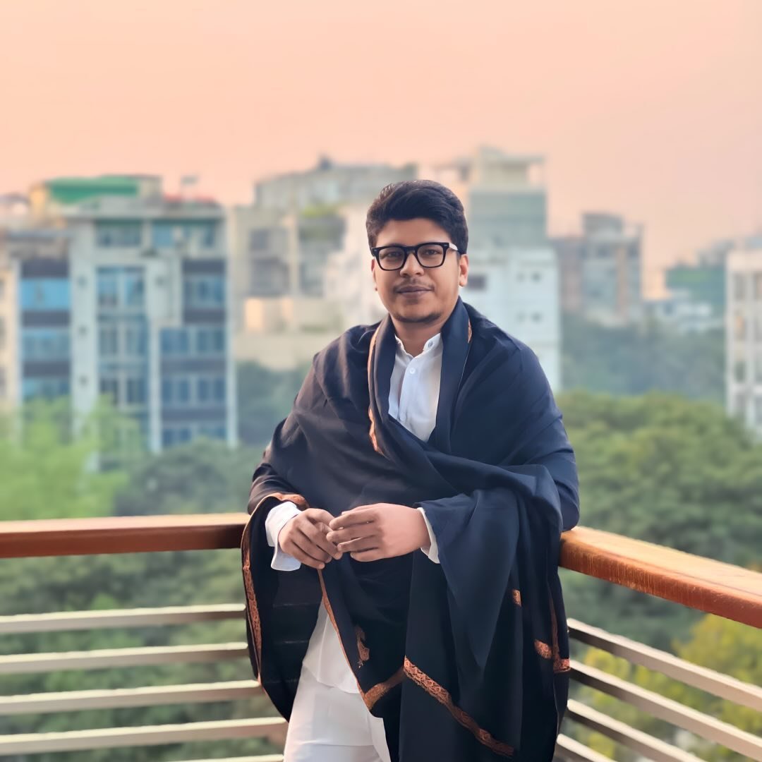 Raihan Khan wrapped in a dark shawl over a white kurta, posing on a rooftop with the city skyline in the background at dusk.
