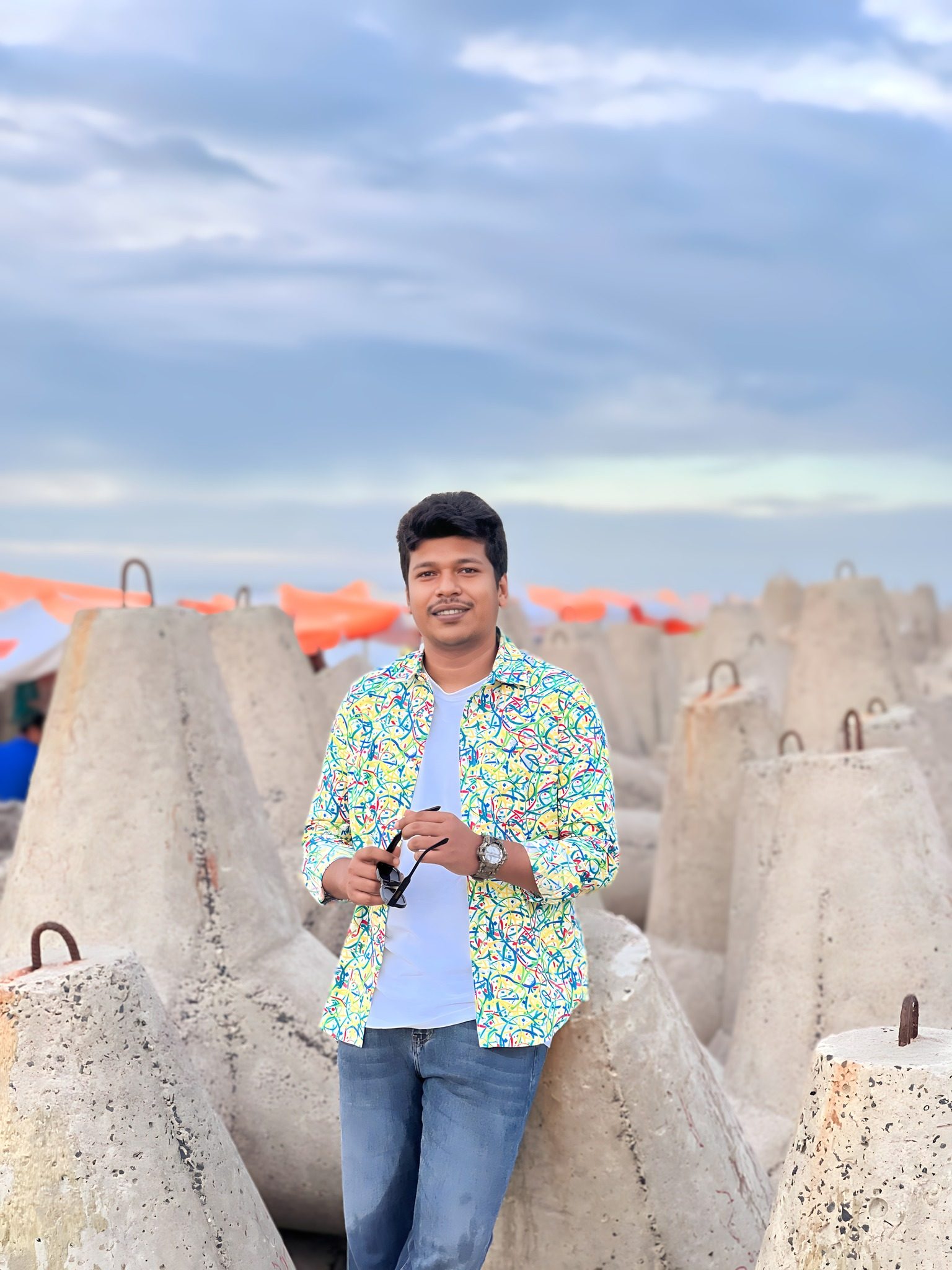 Raihan Khan in a colorful printed shirt stands among concrete breakwater blocks on a beach, holding his sunglasses.