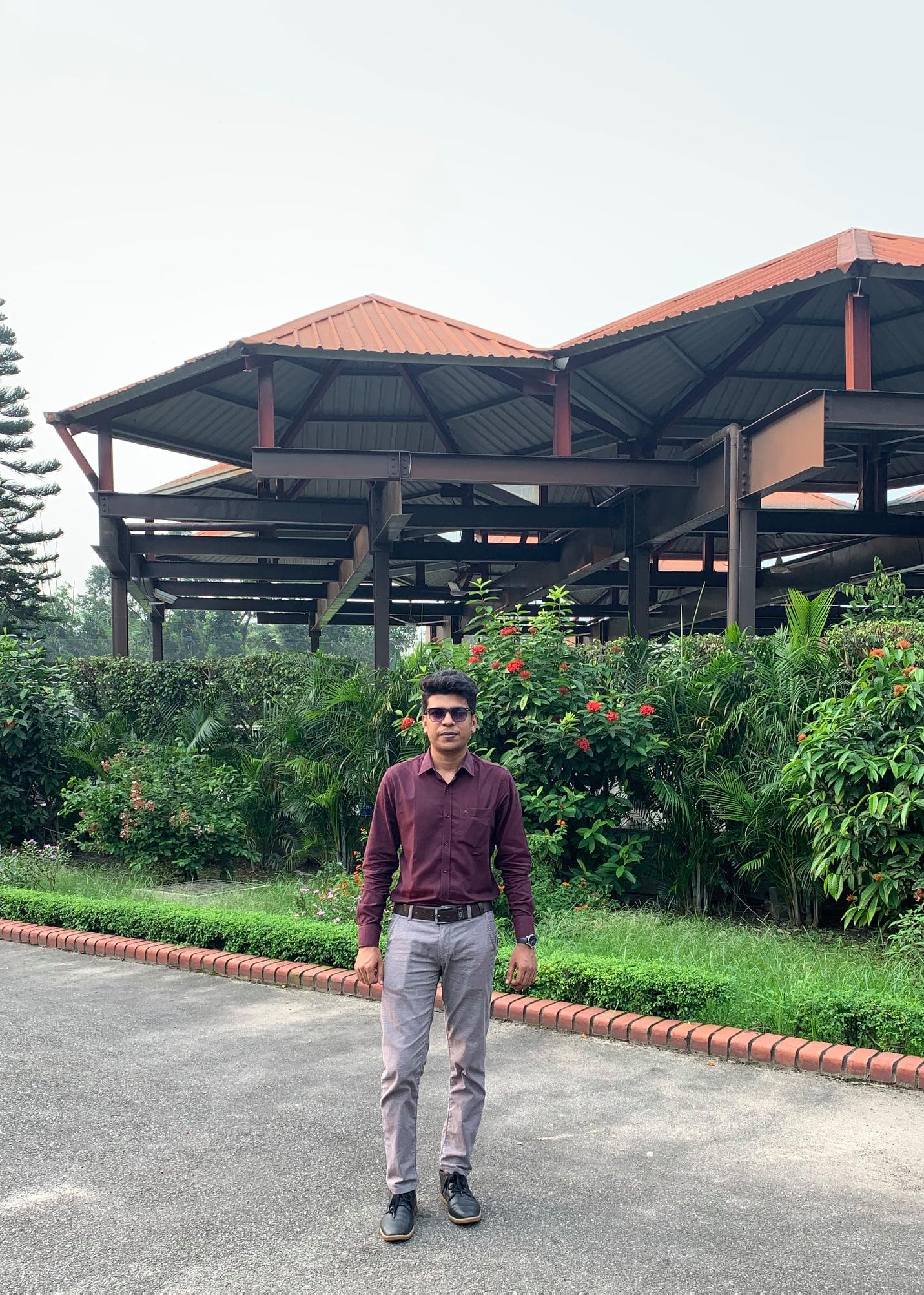 Raihan Khan standing in a landscaped garden pavilion with a red tin roof in Bangladesh.