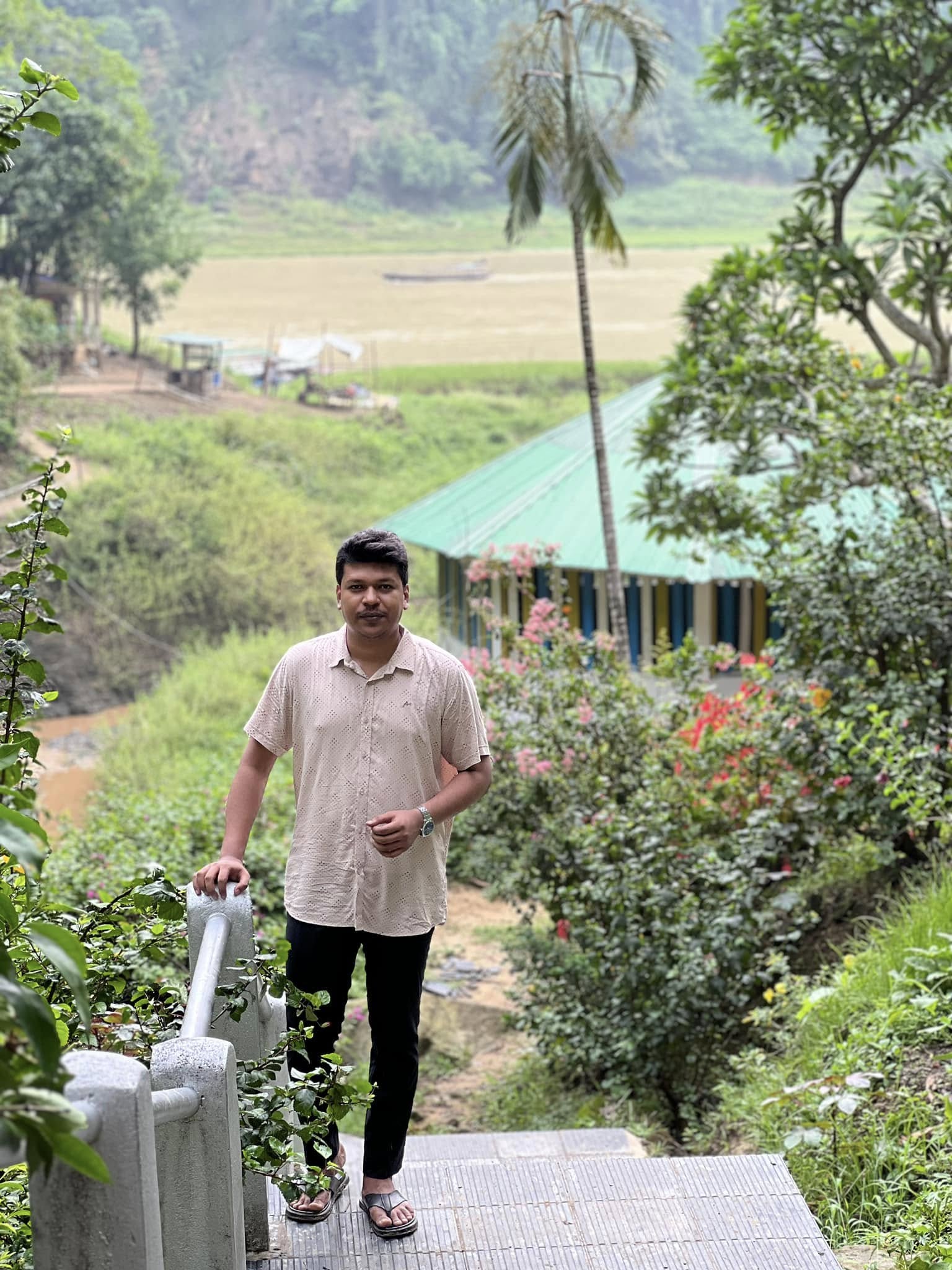 Raihan Khan pausing on a hillside staircase overlooking a river and a green-roofed cottage.