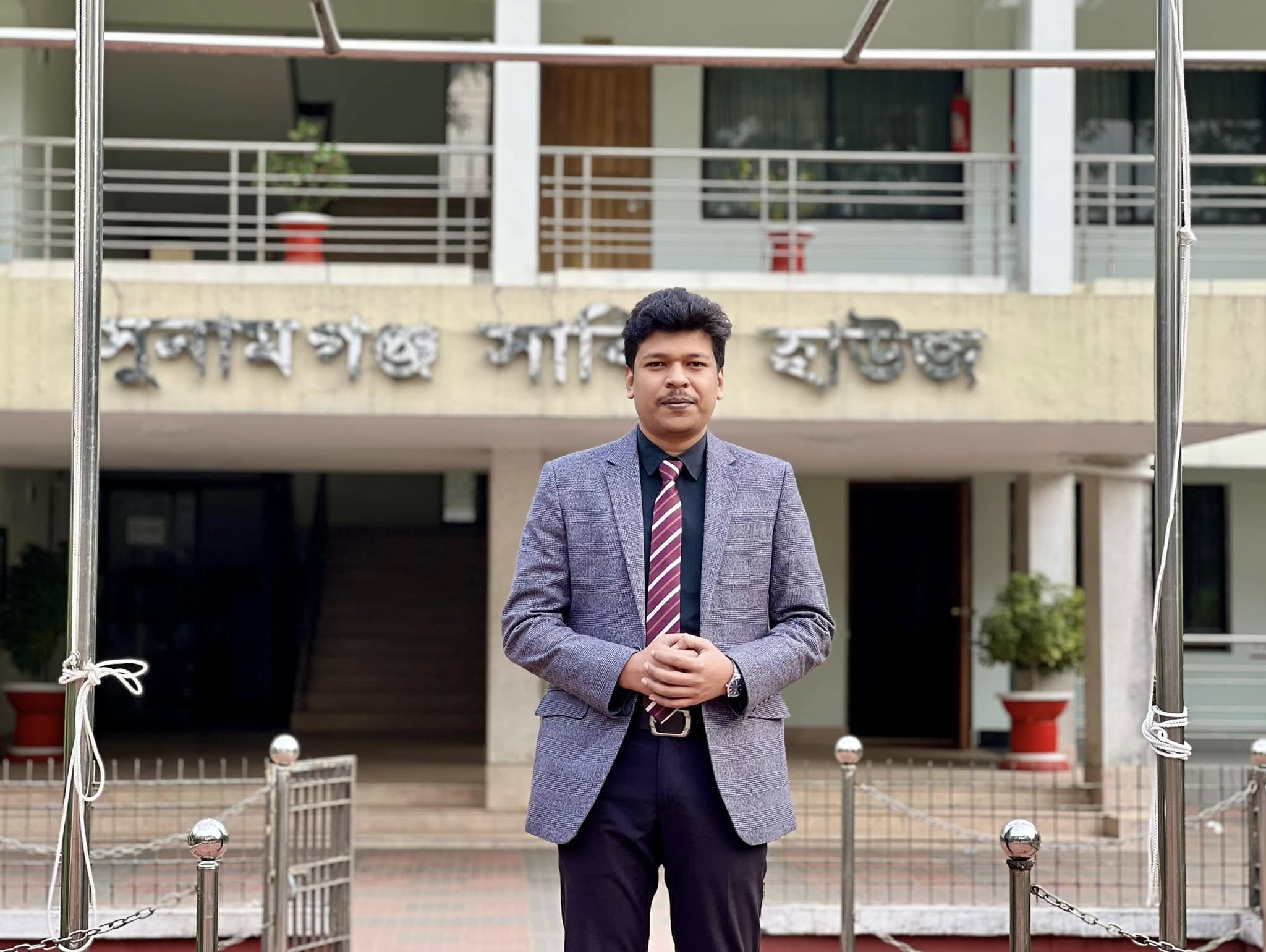 Raihan Khan in a blazer and tie standing before a historic memorial building with Bengali signage.