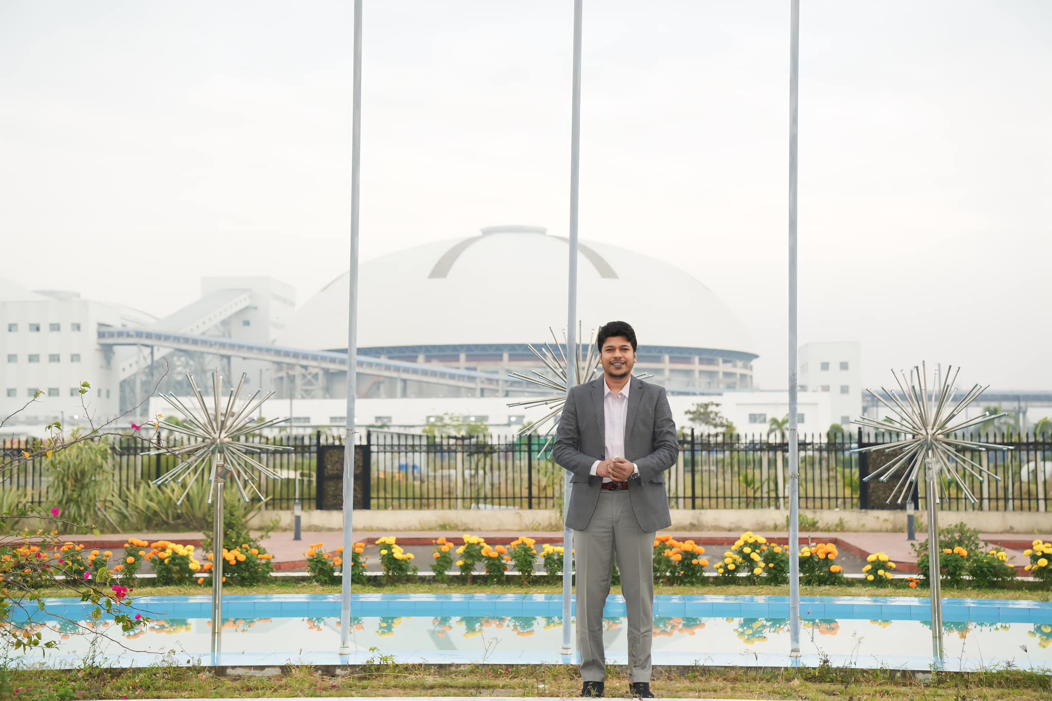 Raihan Khan standing near flagpoles and starburst sculptures with a large dome building in the background.