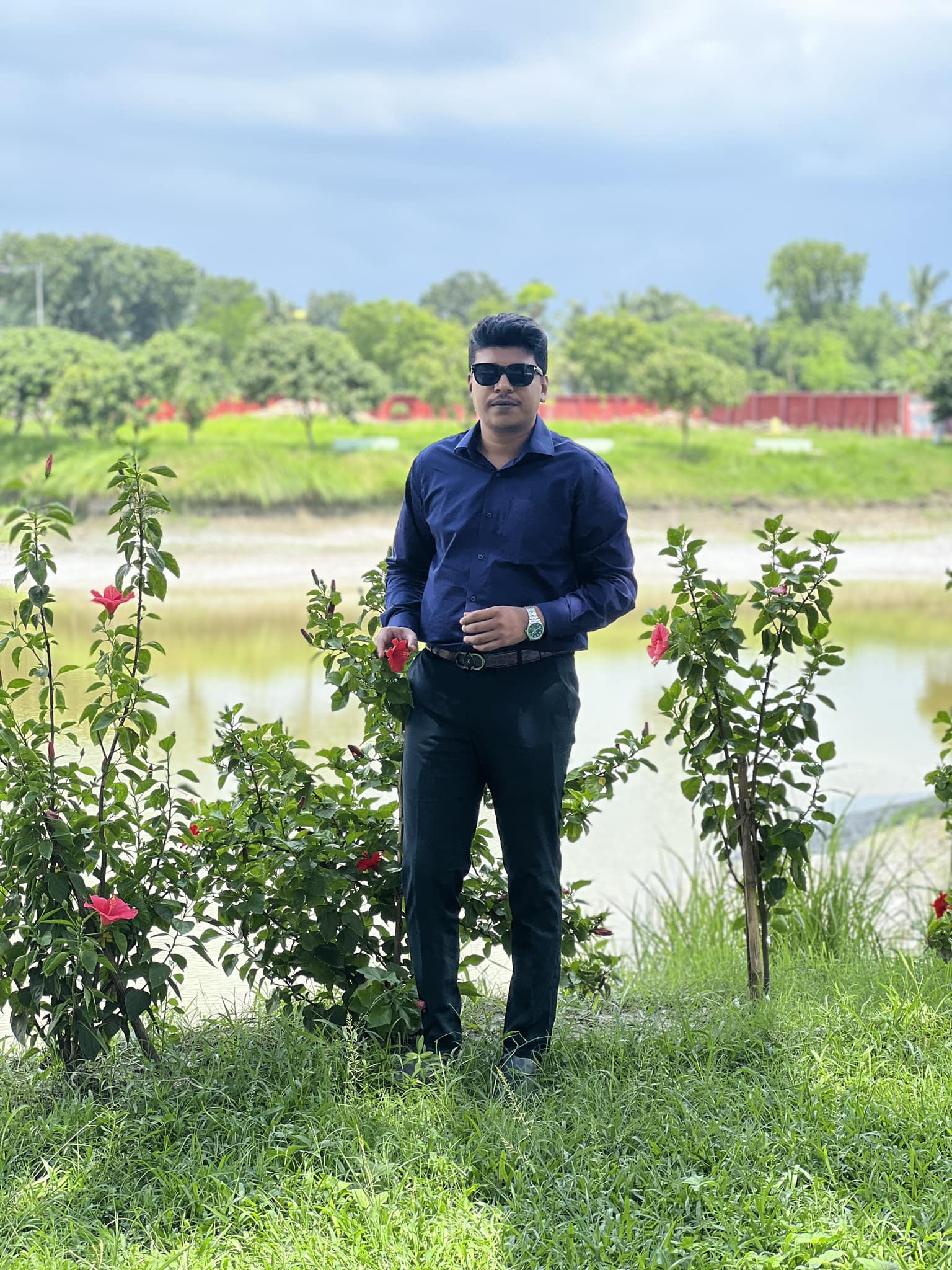 Raihan Khan posing beside blooming red hibiscus bushes along a riverbank in Bangladesh.