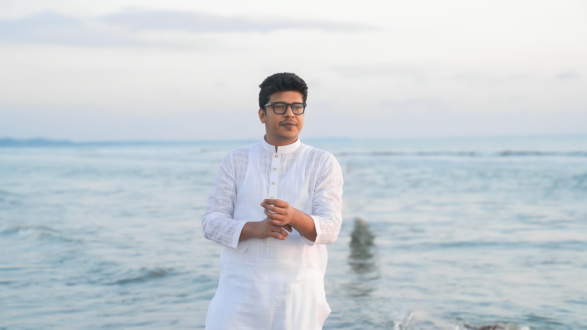 Raihan Khan in a white kurta stands by the sea, gazing into the distance with calm waves in the background.