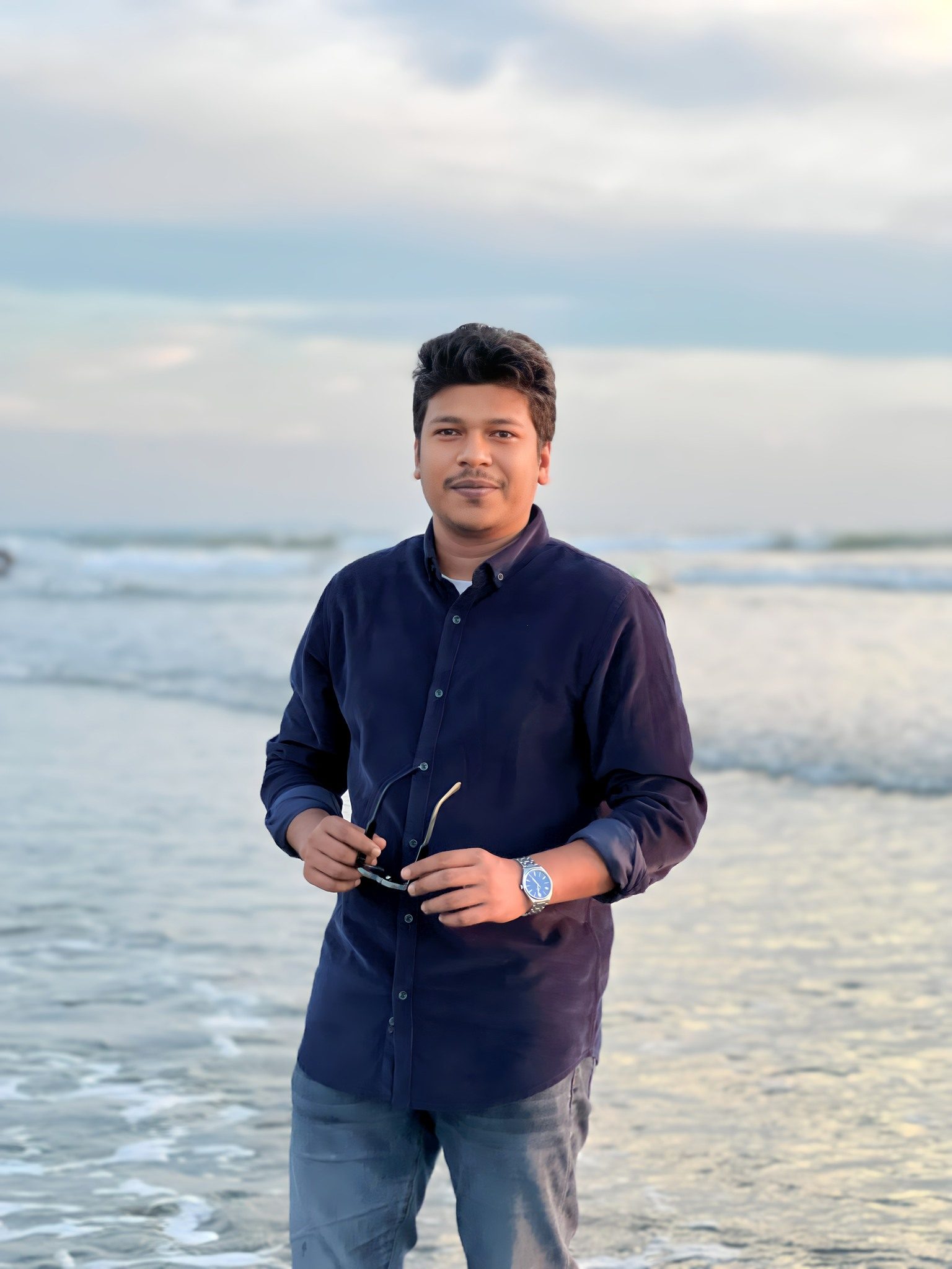 Raihan Khan in a navy shirt stands on the shoreline in the evening, holding his sunglasses as gentle waves roll in.