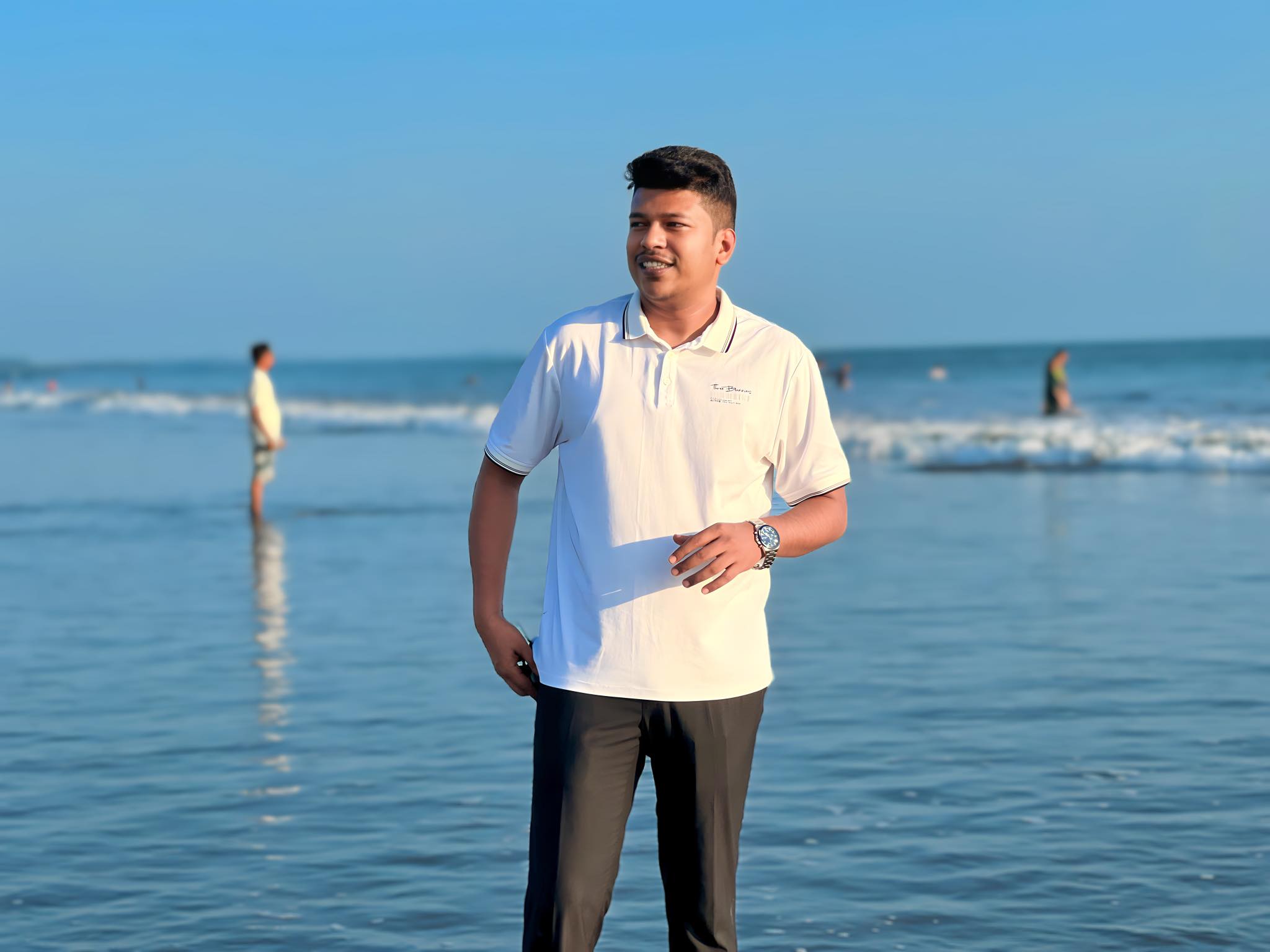 Raihan Khan in a white polo shirt smiles while standing on a sunny beach shoreline with waves and other visitors in the background.