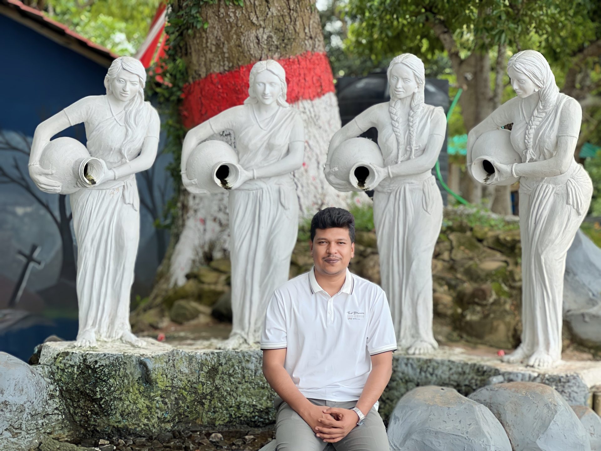 Raihan Khan sits before four statues of women carrying water jugs at a park, with a large tree trunk marked in red and white behind him.