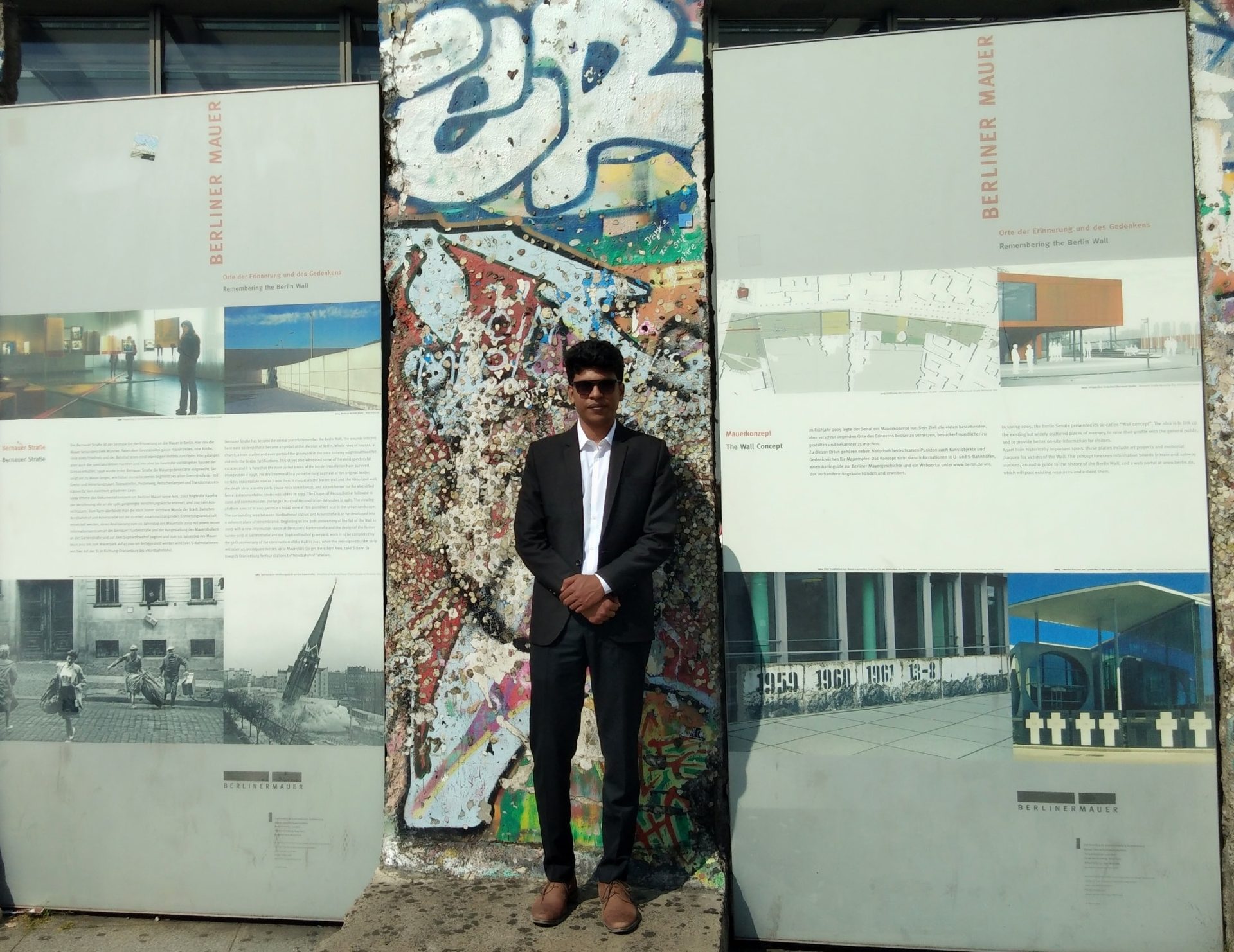 Raihan Khan standing beside a graffiti-covered remnant of the Berlin Wall on Bernauer Strasse.