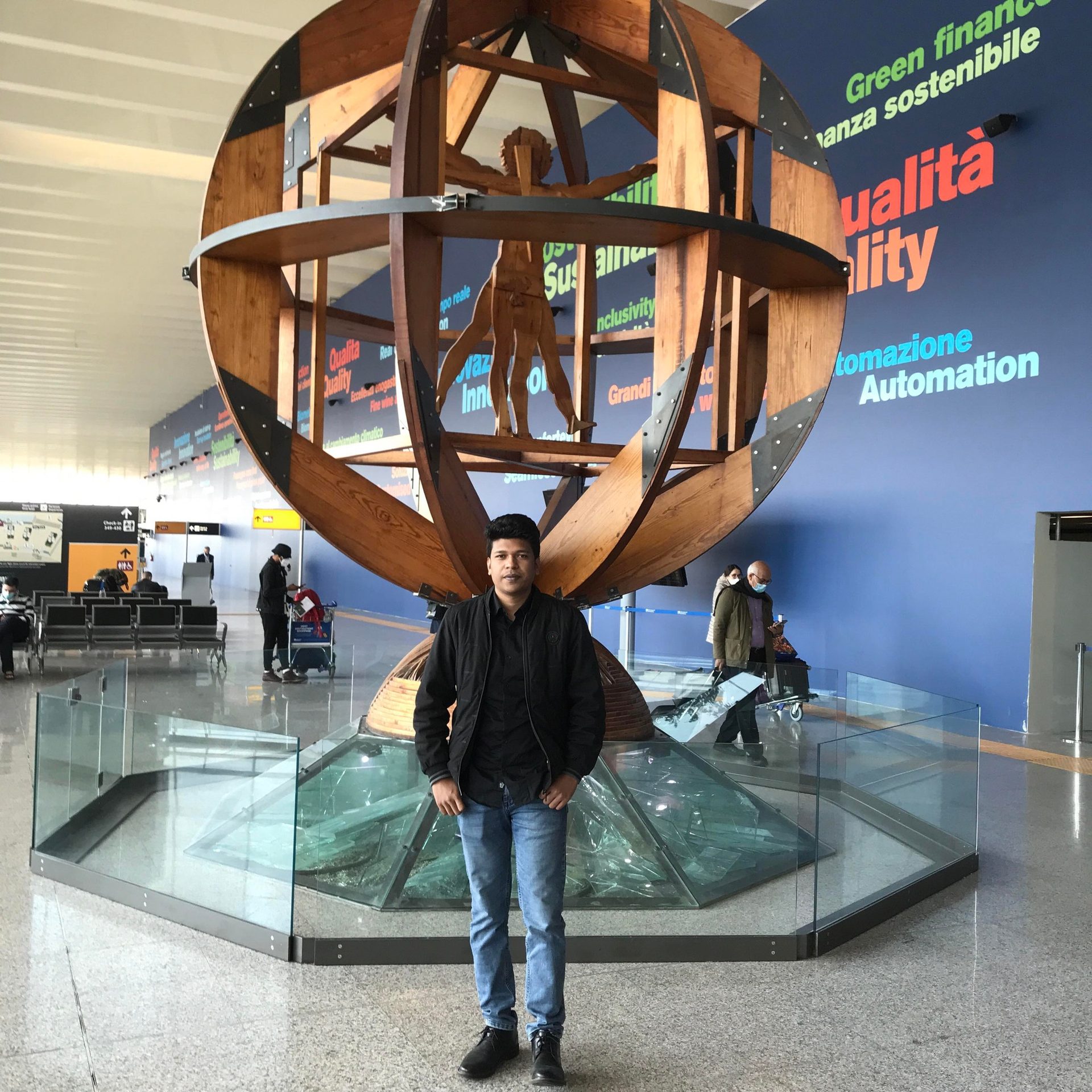 Raihan Khan standing beneath a large wooden Vitruvian Man sculpture inside Rome's Fiumicino Airport.
