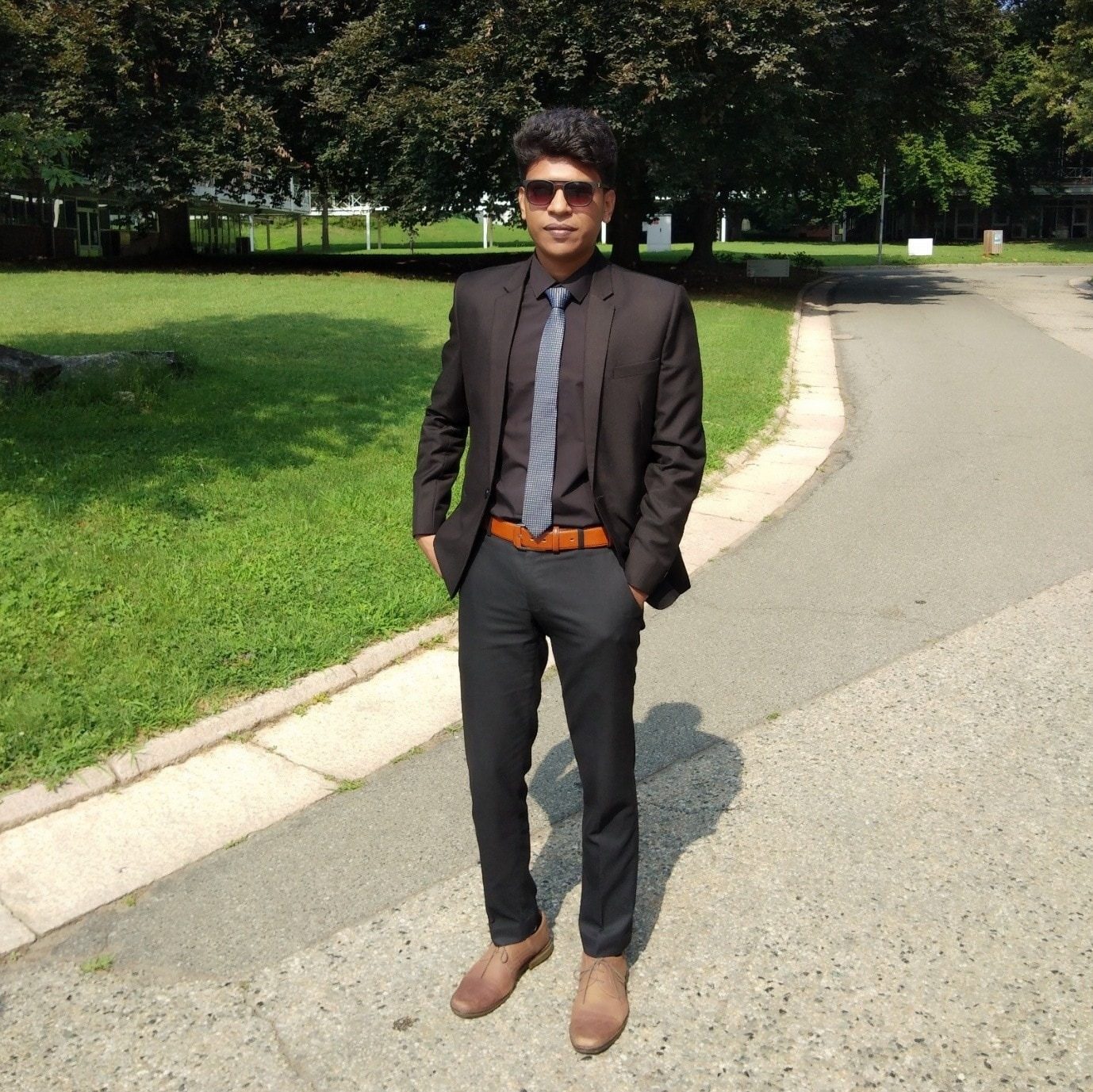 Raihan Khan posing in a suit and sunglasses along a tree-lined pathway in a European park.