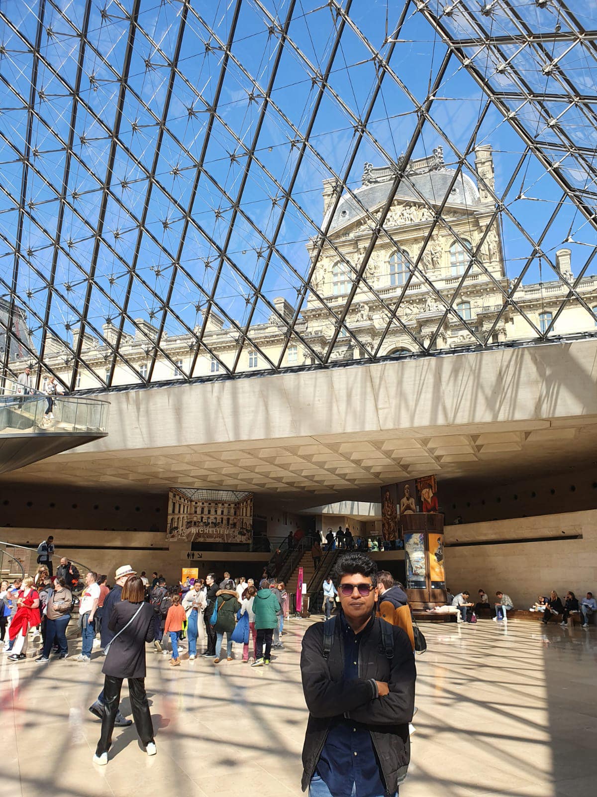Raihan Khan standing inside the glass pyramid entrance hall of the Louvre Museum in Paris.
