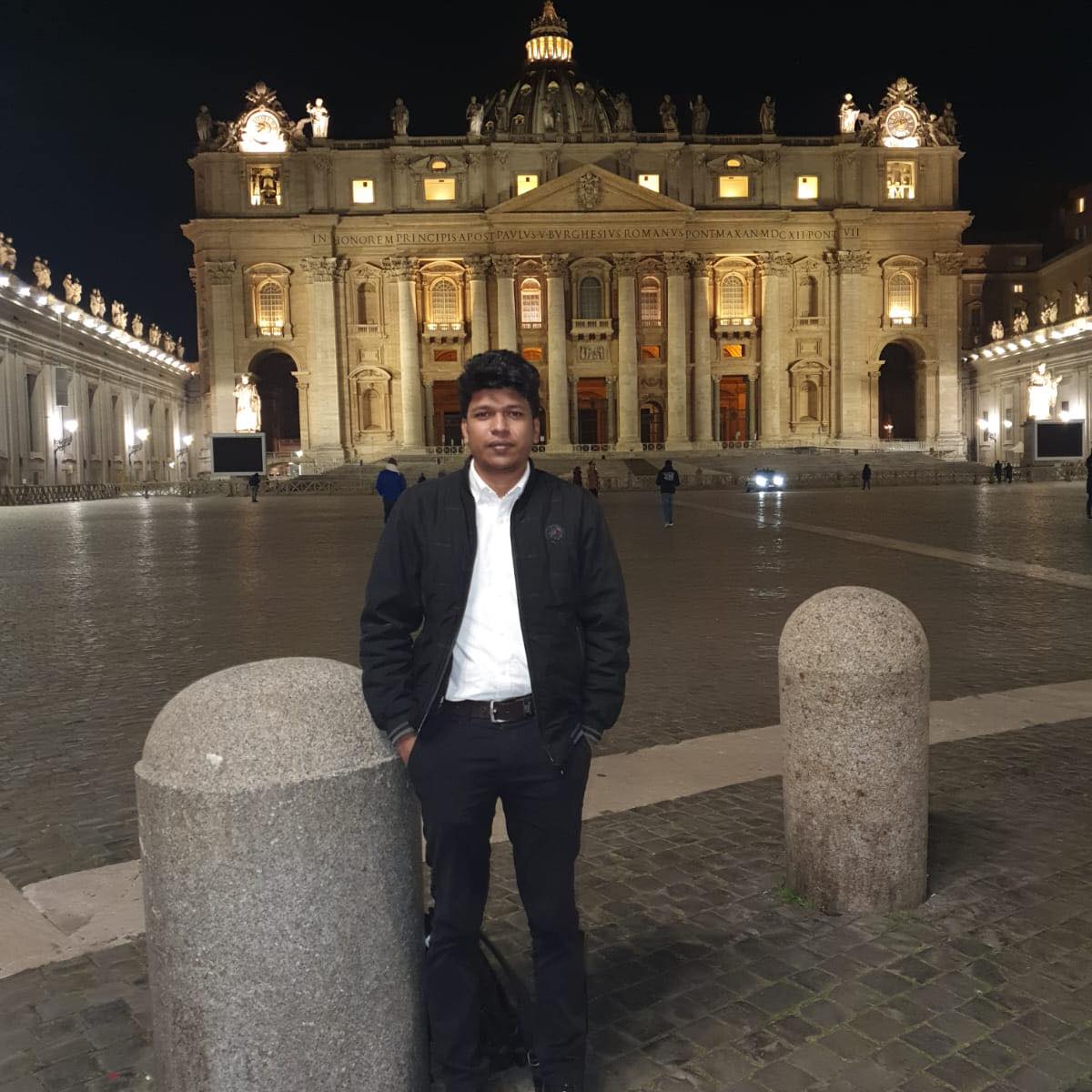Raihan Khan standing alone in St. Peter's Square with the illuminated basilica behind him at night.