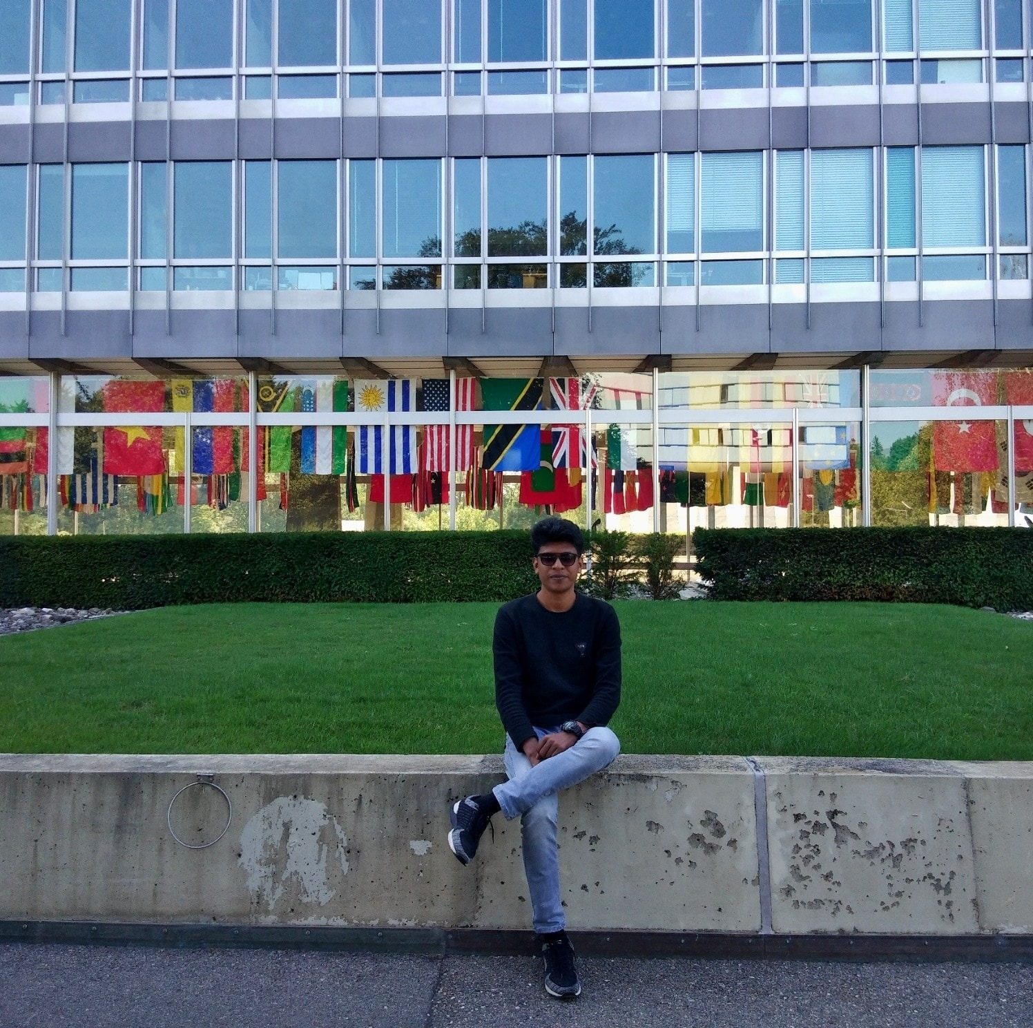 Raihan Khan sitting on a wall in front of the United Nations building in Geneva, flanked by national flags.