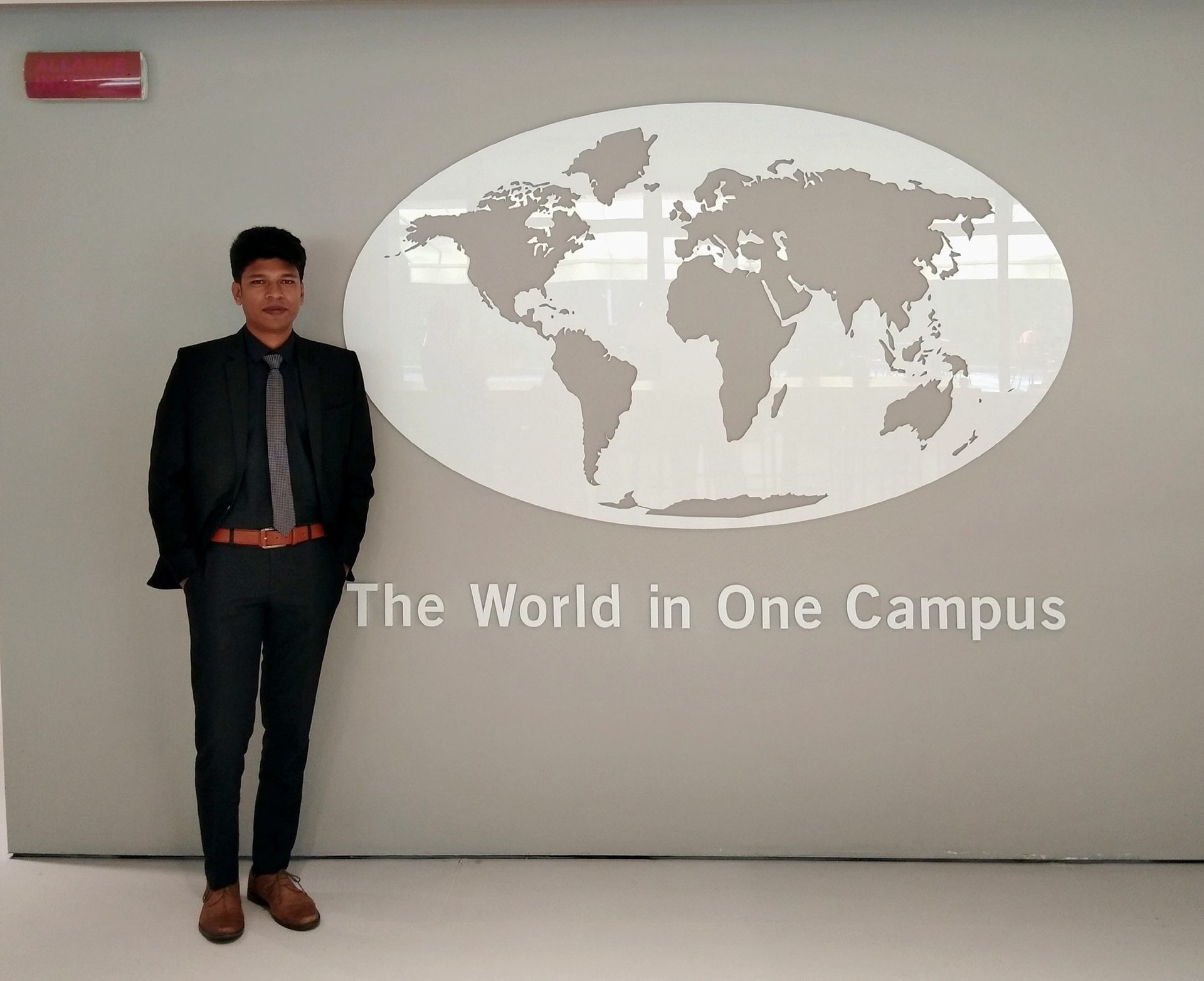 Raihan Khan standing beside a wall-mounted world map reading 'The World in One Campus'.